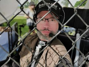 Photographie de Jonathan Marchand, un homme en fauteuil roulant électrique avec une barbe, une casquette, derrière un grillage.