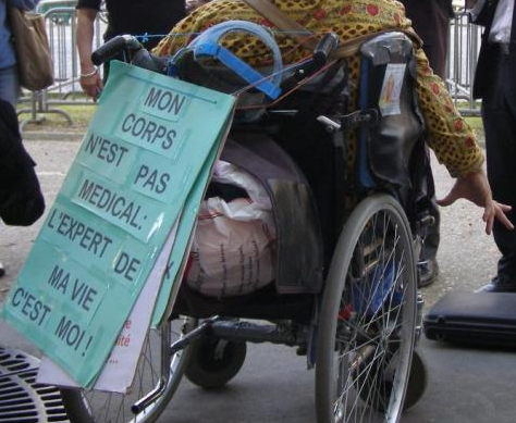 Photographie de dos d'une personne en fauteuil roulant manuel pendant une manifestation. Une pancarte est accrochée au fauteuil : "Mon corps n'est pas médical : l'expert de ma vie c'est moi"